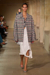 Model walks a runway in an oversized plaid jacket over a white draped dress, paired with gray heeled sandals.