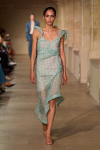 Model walking the runway in a pale mint green, semi-sheer dress with ruffled shoulder straps and a wrapped, tiered skirt.