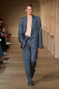 Model walks a fashion runway in an oversized blue suit with a pale pink blouse and a dark brown belt, mid-stride.