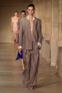 Model walks the runway in a taupe oversized suit with white-collar shirt and cuffs, hands in pockets.