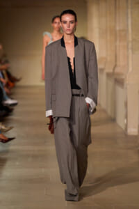 Model walks the runway in a loose gray suit with an oversized blazer, black top, and wide trousers, holding a brown clutch.