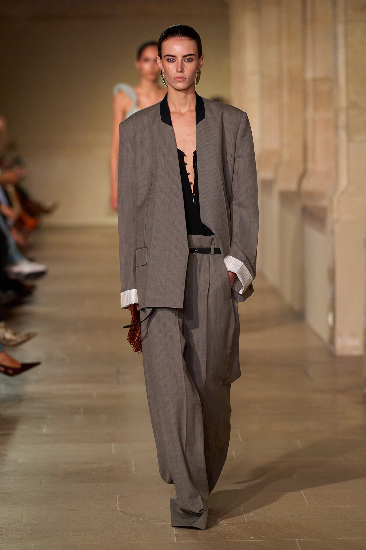 Model walks the runway in a loose gray suit with an oversized blazer, black top, and wide trousers, holding a brown clutch.