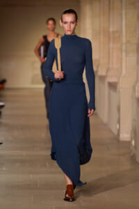 Model walks the runway in a navy textured knit turtleneck dress, carrying a tan shoulder bag and wearing brown loafers.