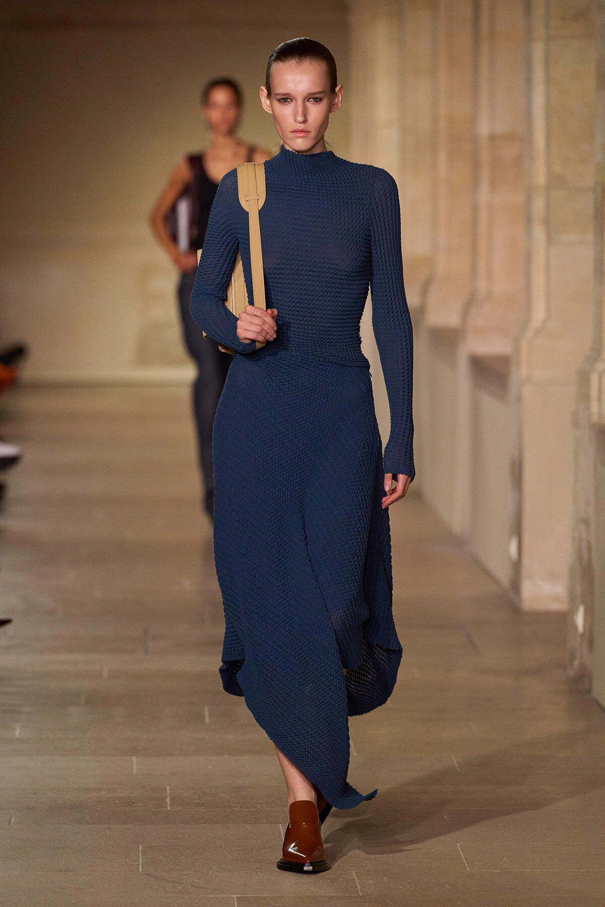 Model walks the runway in a navy textured knit turtleneck dress, carrying a tan shoulder bag and wearing brown loafers.