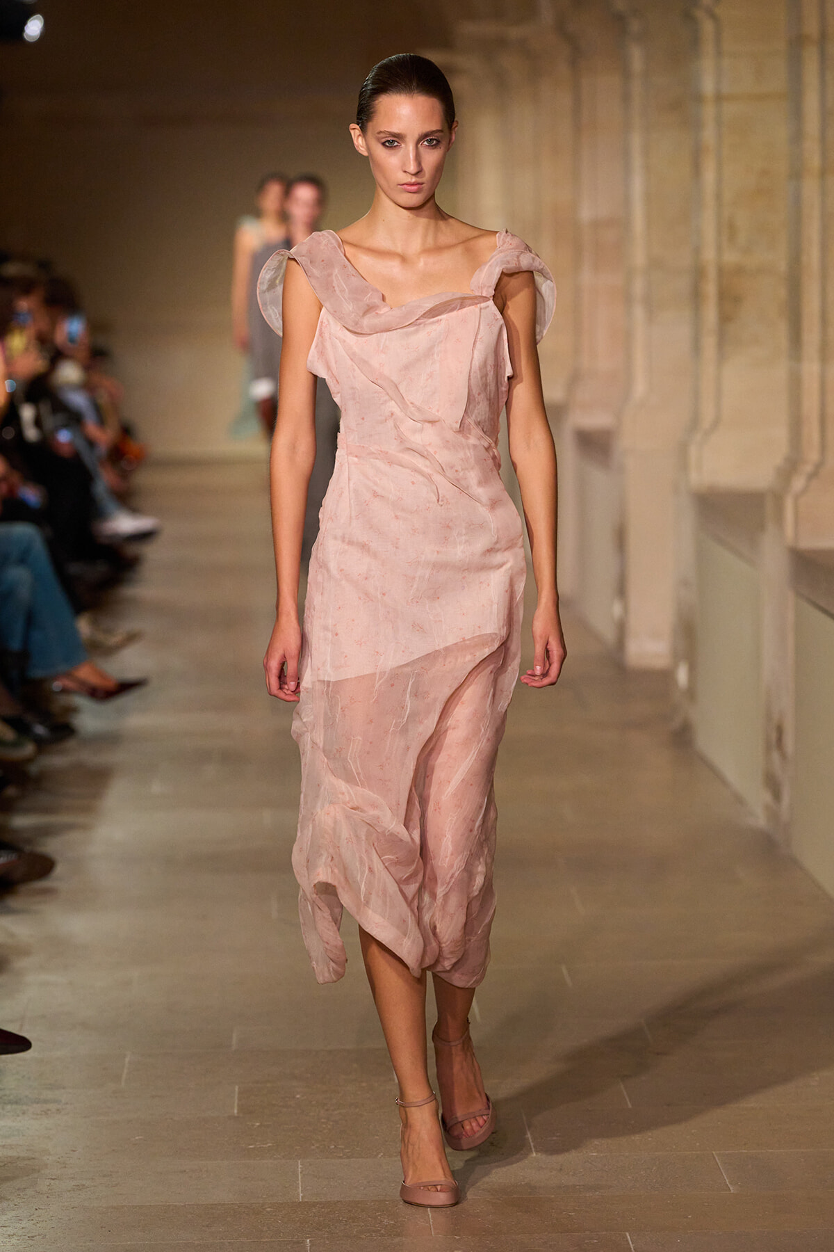 Model walks a fashion runway in a pale pink draped midi dress with a soft off-shoulder neckline and nude sandals