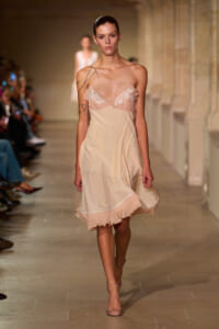 Model walking a runway in a pale peach slip dress with lace bust and thin straps, featuring a ruffled hem.