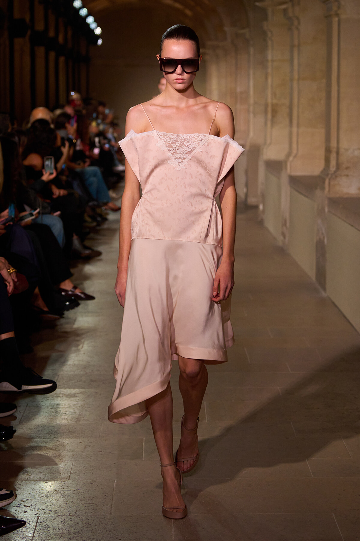 Model on a fashion runway wearing a pale pink off-the-shoulder lace blouse and a satin high-low skirt, with oversized sunglasses.