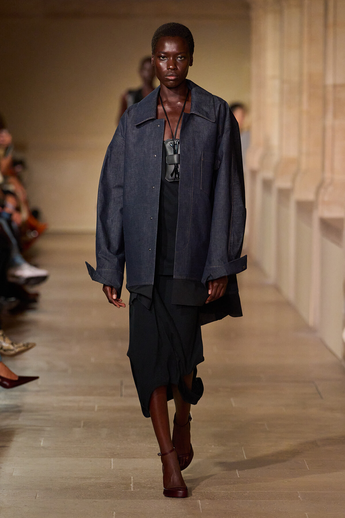 Model on a runway wearing an oversized dark denim jacket over a black layered outfit, burgundy heels, and a chunky black pendant necklace.