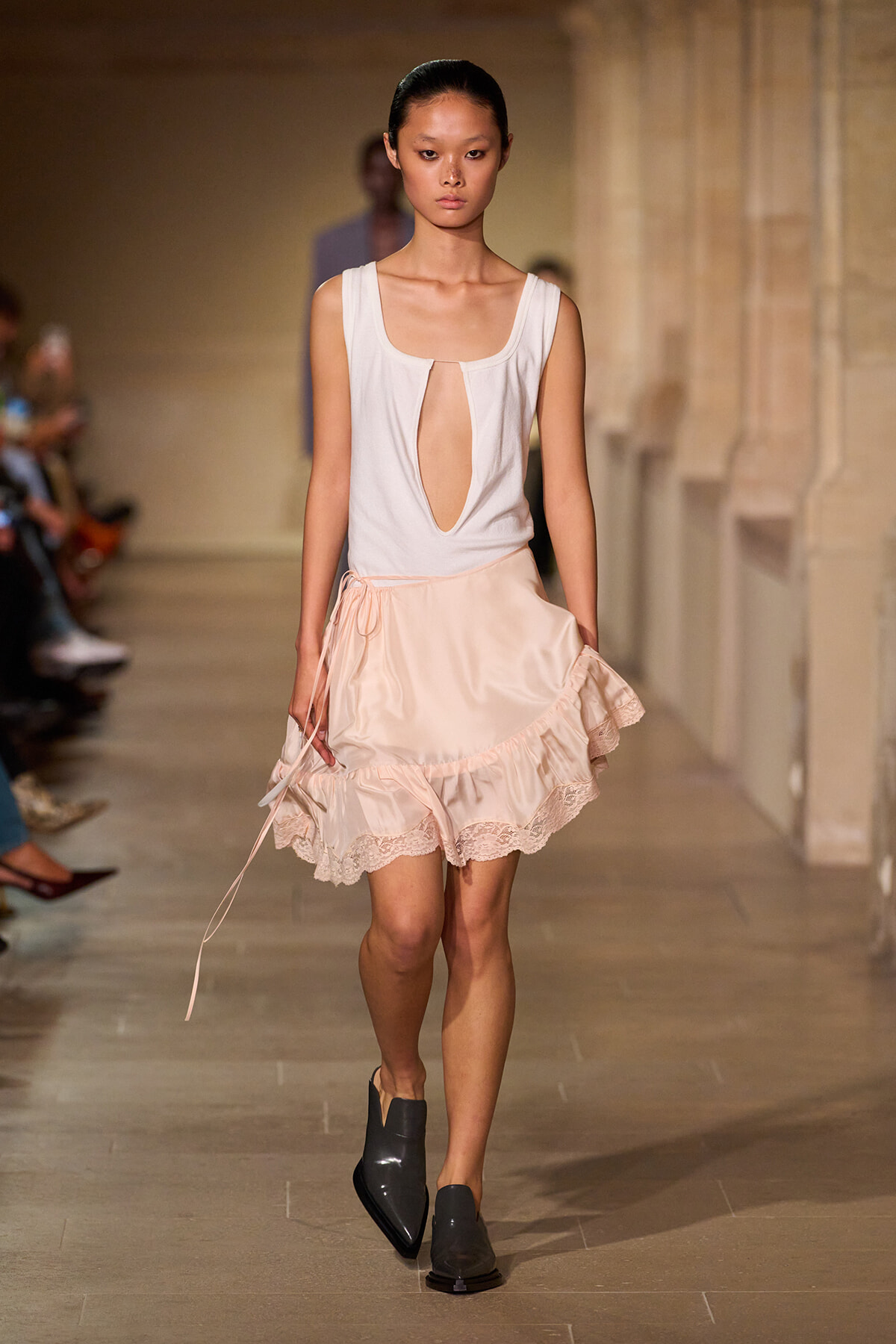 Model walks the runway in a white sleeveless top with a deep keyhole at the center and a pale pink, lace-trimmed, tiered skirt.