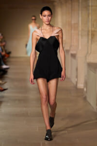 Model walks the runway in a black romper with pale pink feather-like shoulder accents.