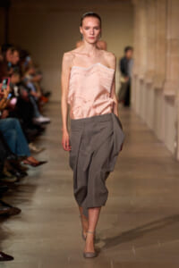 Model walking a fashion runway wearing a pale pink, off-shoulder satin top and gray draped cropped trousers, with minimal jewelry and neutral heels.