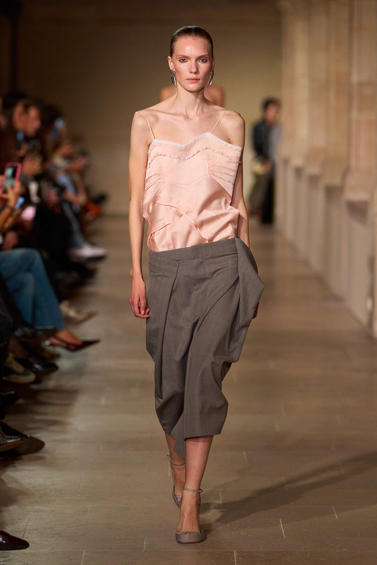 Model walking a fashion runway wearing a pale pink, off-shoulder satin top and gray draped cropped trousers, with minimal jewelry and neutral heels.