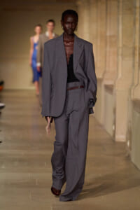 Model on a fashion runway wearing an oversized gray suit with a black top, brown belt, and a small clutch.