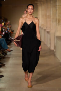 Model walks the runway in a black halter dress with wide, draped trousers-like pants, carrying a burgundy handbag under her arm.