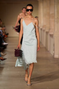 Model walks the runway in a pale blue slip dress with a lace hem, oversized sunglasses, and a burgundy stacked clutch.