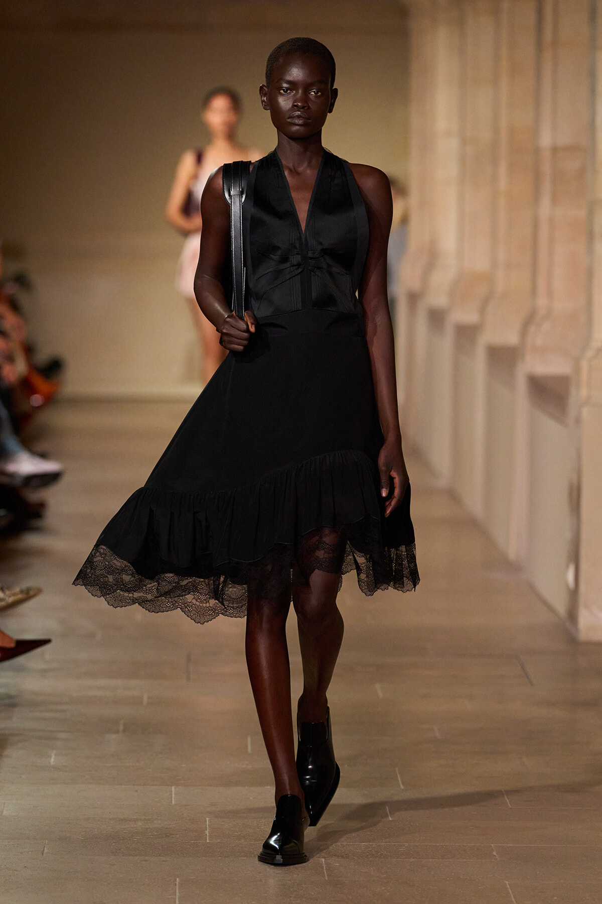 Model walking a runway in a knee‑length black dress with a V‑neck and lace-trimmed hem, carrying a shoulder bag.
