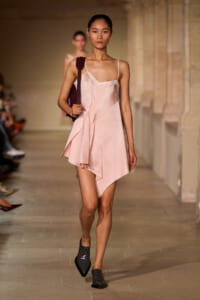 Model walks a runway in a pale pink lace-trim slip dress with an asymmetric hem, carrying a burgundy shoulder bag.