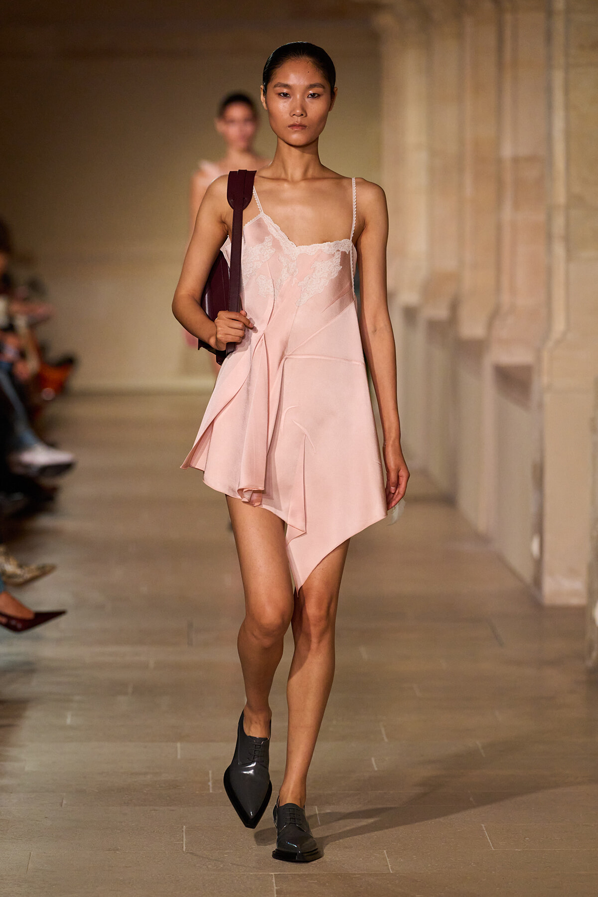 Model walks a runway in a pale pink lace-trim slip dress with an asymmetric hem, carrying a burgundy shoulder bag.