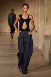 Model on a runway wearing a black sleeveless cut-out top and baggy dark denim jeans, carrying a burgundy handbag over her shoulder.