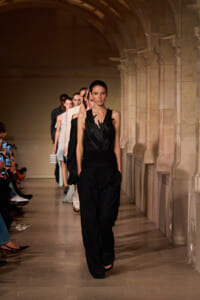 Model walks the runway in a black sleeveless jumpsuit as a line of models follows in a stone-arched corridor fashion show.