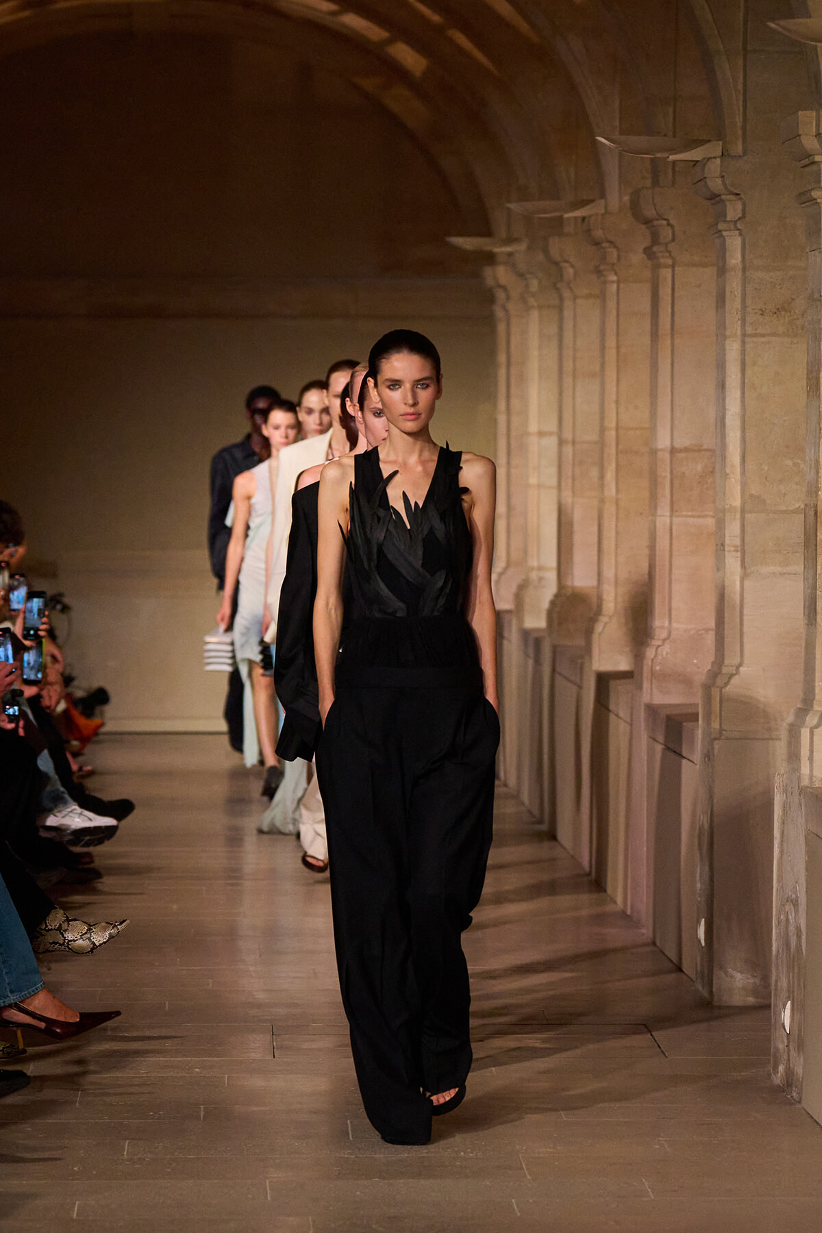 Model walks the runway in a black sleeveless jumpsuit as a line of models follows in a stone-arched corridor fashion show.