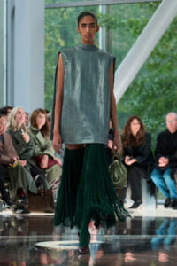 Model runway look: metallic silver sleeveless tunic with cape-like sleeves and a long green fringe skirt, carrying a green handbag, audience in background