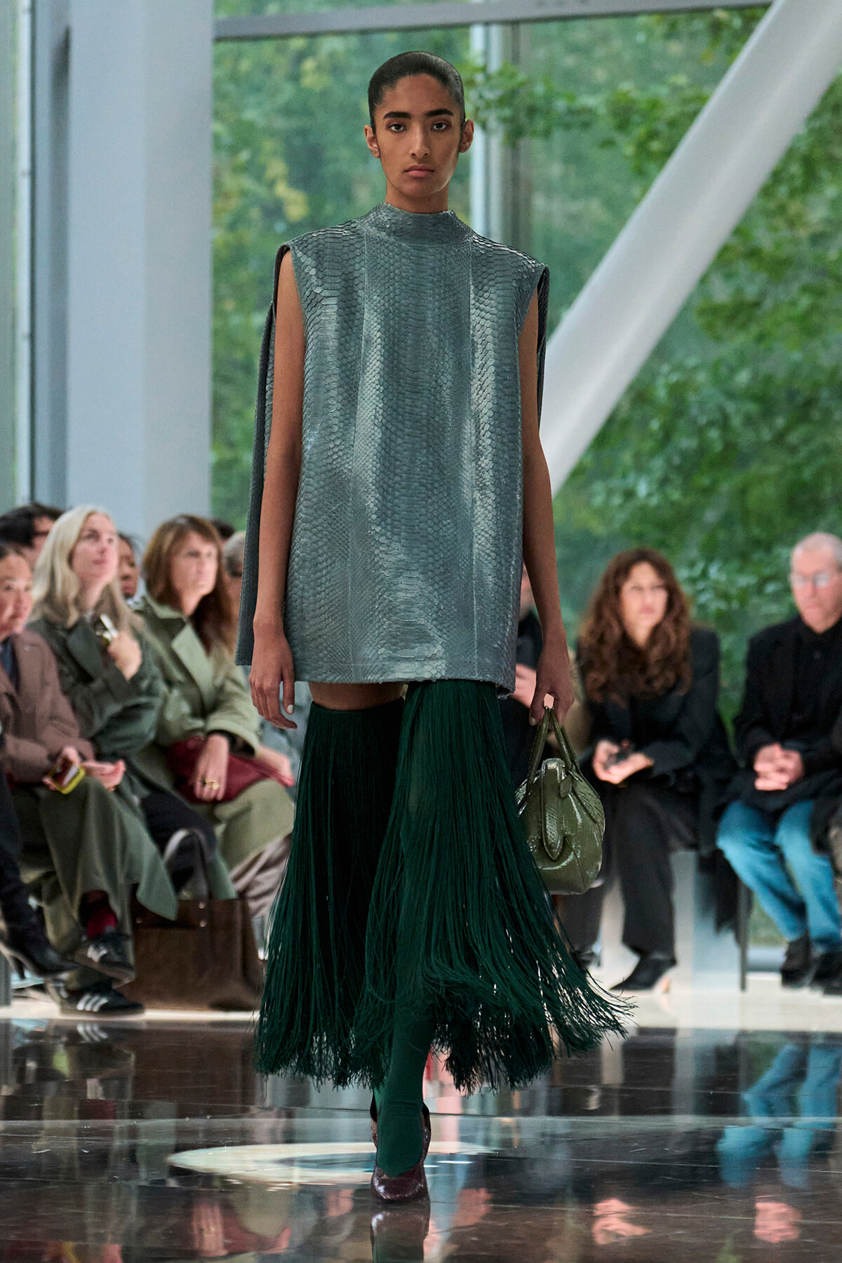 Model runway look: metallic silver sleeveless tunic with cape-like sleeves and a long green fringe skirt, carrying a green handbag, audience in background