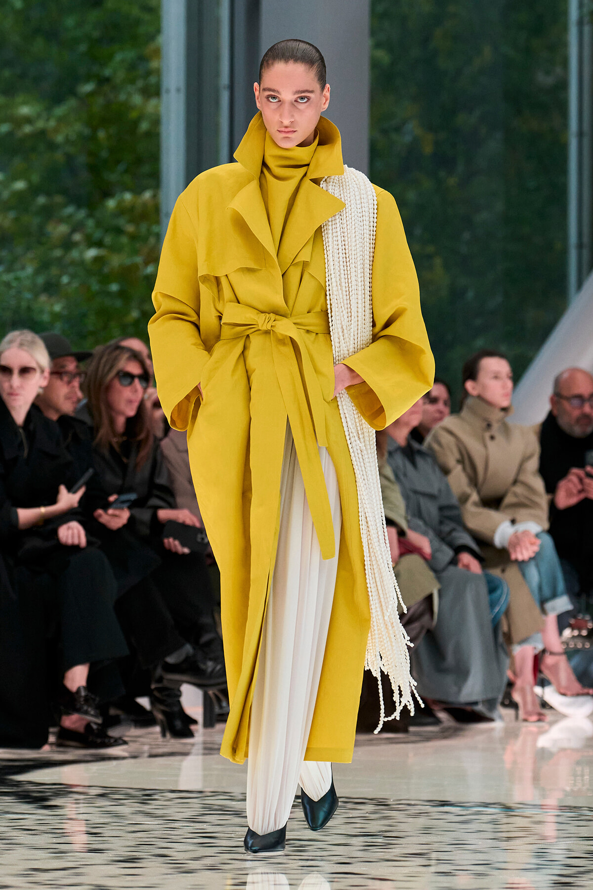 Model in a mustard-yellow belted trench coat with white pleated pants and a long cream scarf, walking a glossy runway at a fashion show.