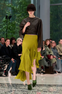 Model walking a fashion runway in a dark cropped leather top and lime-green pleated high-low skirt; audience seated in background.