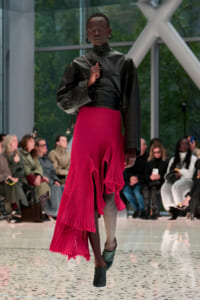 Model walking a fashion runway in a black leather top and a bright pink asymmetrical pleated skirt, with audience in the background.