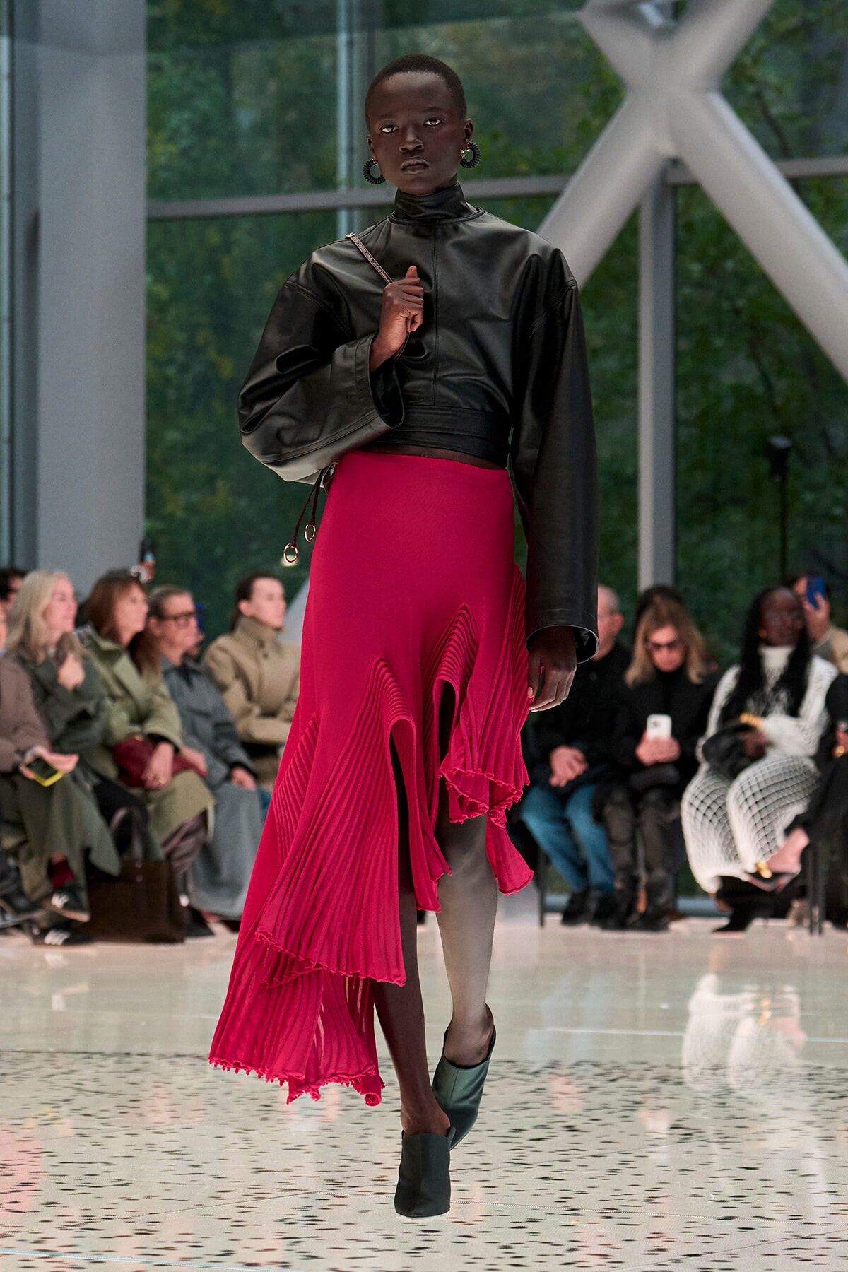 Model walking a fashion runway in a black leather top and a bright pink asymmetrical pleated skirt, with audience in the background.