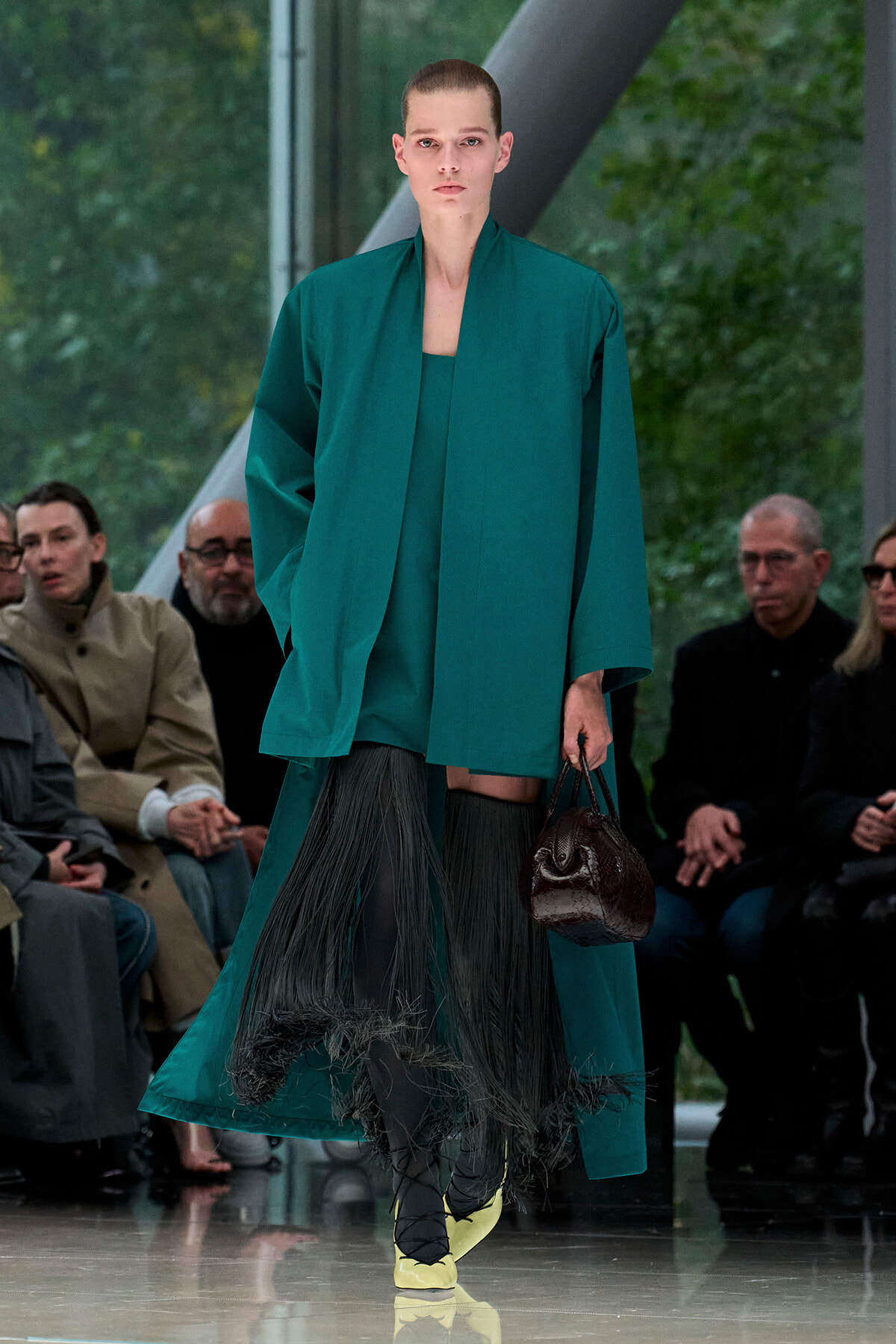 Model walking a fashion show in a teal coat over a fringe skirt, carrying a dark brown textured bag; yellow shoes visible.