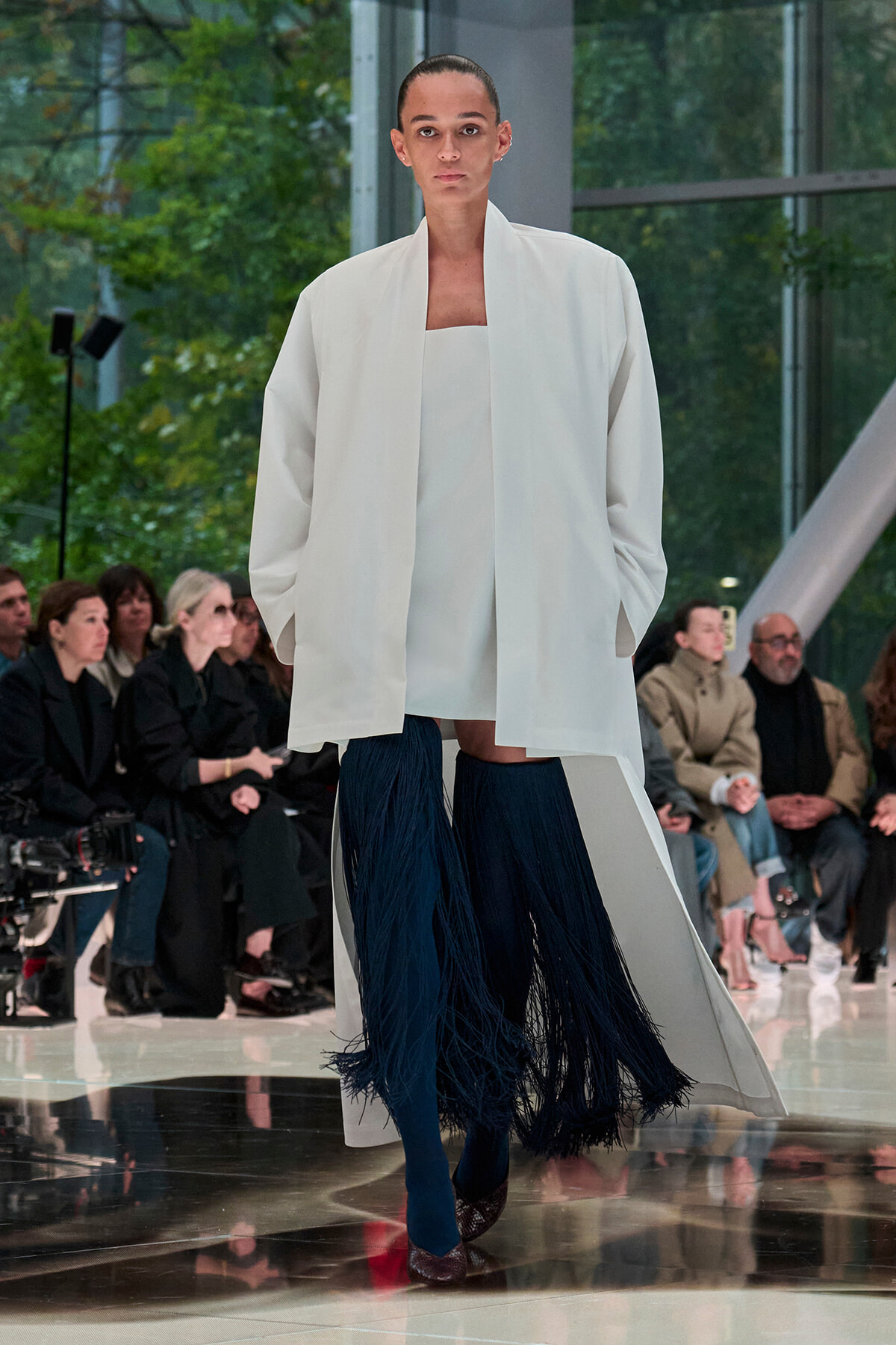 Model walks the runway in an oversized white coat over a white top, paired with dark blue fringe skirt leggings and heels, as audience watches.