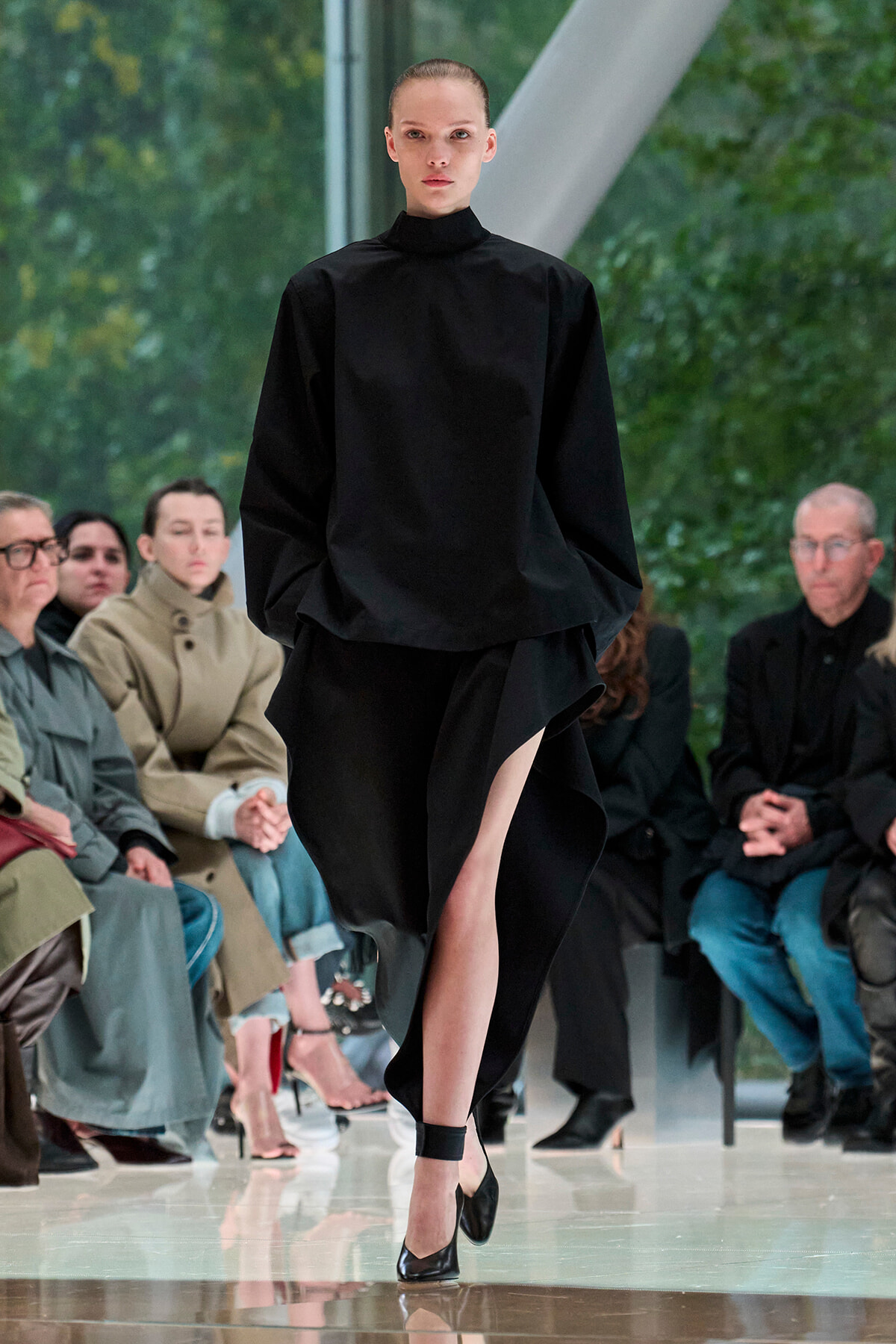 Model on a runway wearing a black high-neck top and draped skirt; seated audience watches in the background.