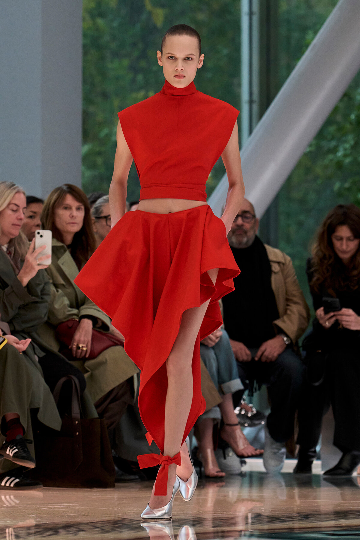 Model walking a runway in a bright red sleeveless crop top and asymmetric high-low skirt; silver heels visible.