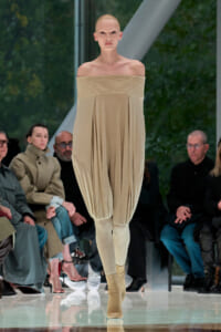 Model on a runway wearing a beige off-the-shoulder pleated top with roomy, draped pants and beige boots; spectators sit along the catwalk.] ,