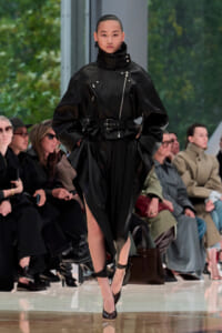 Model strides down runway in a black leather oversized jacket with high collar, belted waist, and matching slit skirt; audience seated along the catwalk