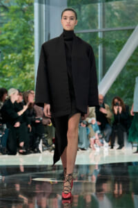 Model walks a runway in a loose black coat over a black turtleneck, paired with red lace-up heels; audience seated along the catwalk in background.