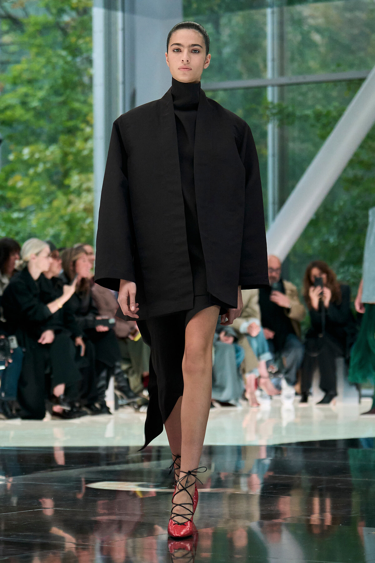 Model walks a runway in a loose black coat over a black turtleneck, paired with red lace-up heels; audience seated along the catwalk in background.