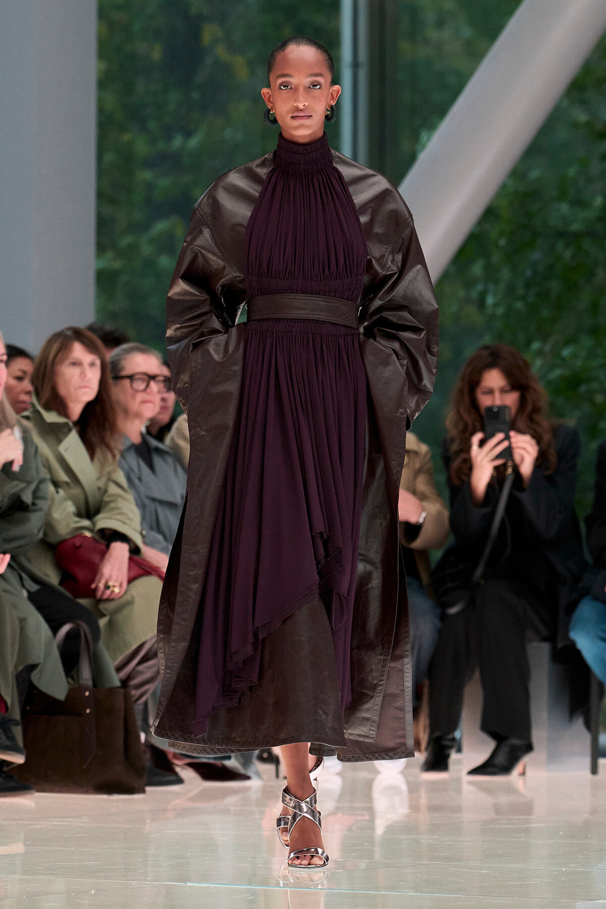Model walking the runway in a dark purple pleated gown with oversized sleeves and a belt, audience in background.
