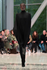 Model on a runway wearing a loose, black high-neck cape dress with draped fabric, black tights, and ankle boots; audience seated along the catwalk