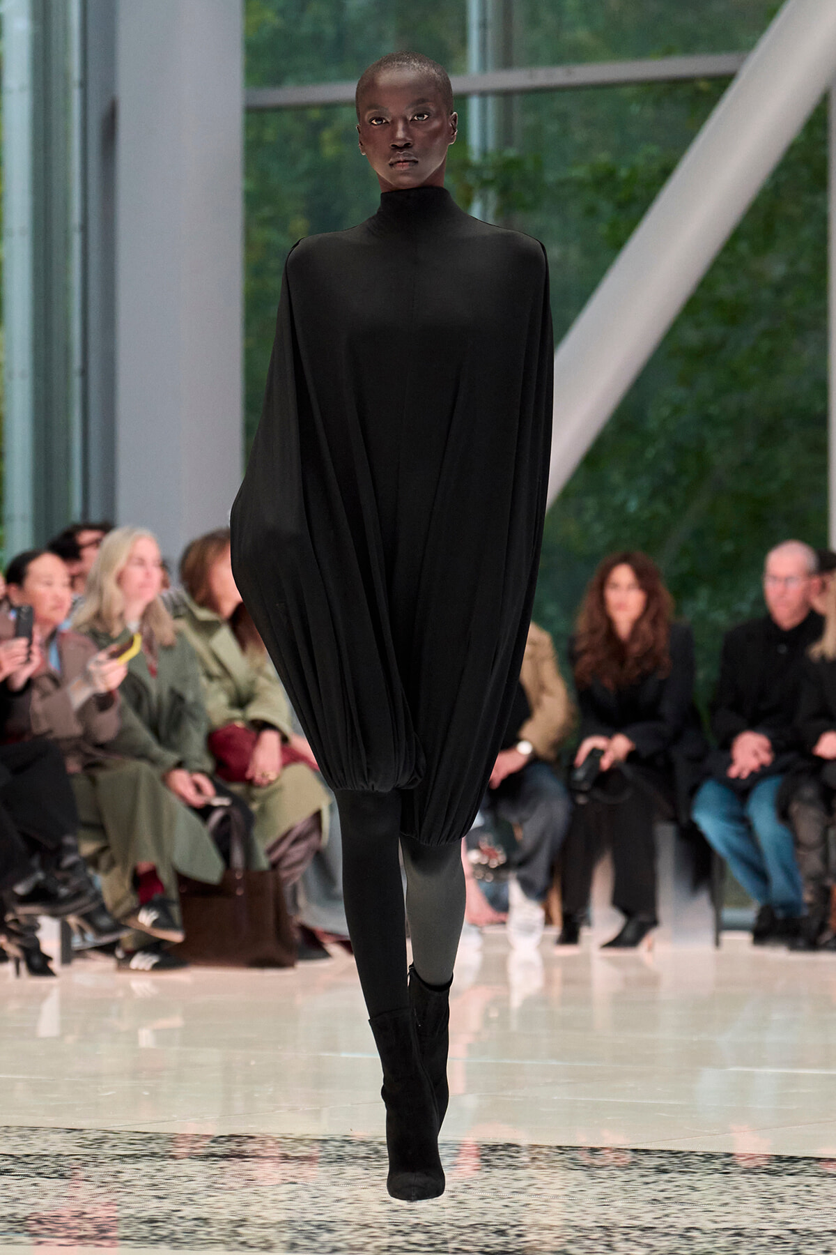 Model on a runway wearing a loose, black high-neck cape dress with draped fabric, black tights, and ankle boots; audience seated along the catwalk