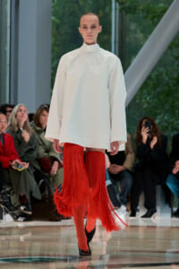 Model walks the runway in a white oversized top with bright red fringe extending below the knees, red tights, and black heels. Runway scene: model in a minimalist white top and dramatic red fringe legwear, audience in background. Runway fashion: a white turtleneck-like top paired with long red fringe leggings and black pumps. A fashion show image of a model wearing a white loose top with red fringed legwear and black heels. White, loose-fitting top with red fringe legwear and black heels showcased on the runway.