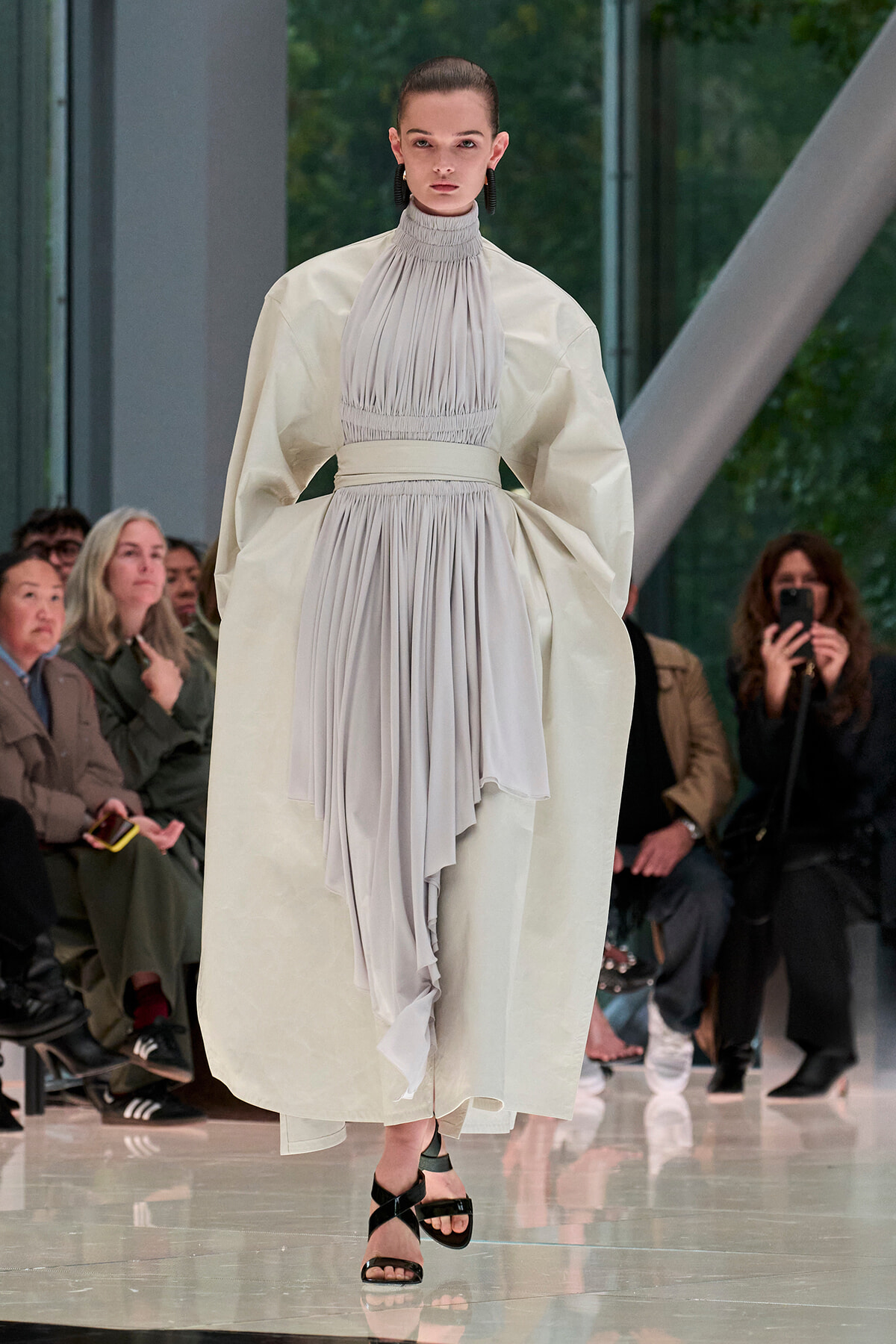 Model walking a fashion runway in a pale, high-neck pleated gown with voluminous cape sleeves and a draped, layered skirt; audience in background.