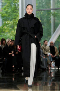 Model in a long black belted coat with a high collar and white pleated trousers, walking a runway while holding a red clutch.