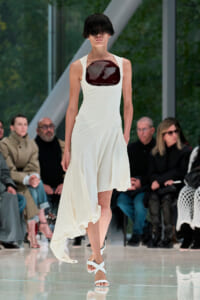 Model walking a runway in a white sleeveless dress with a glossy dark burgundy chest panel, audience in background.