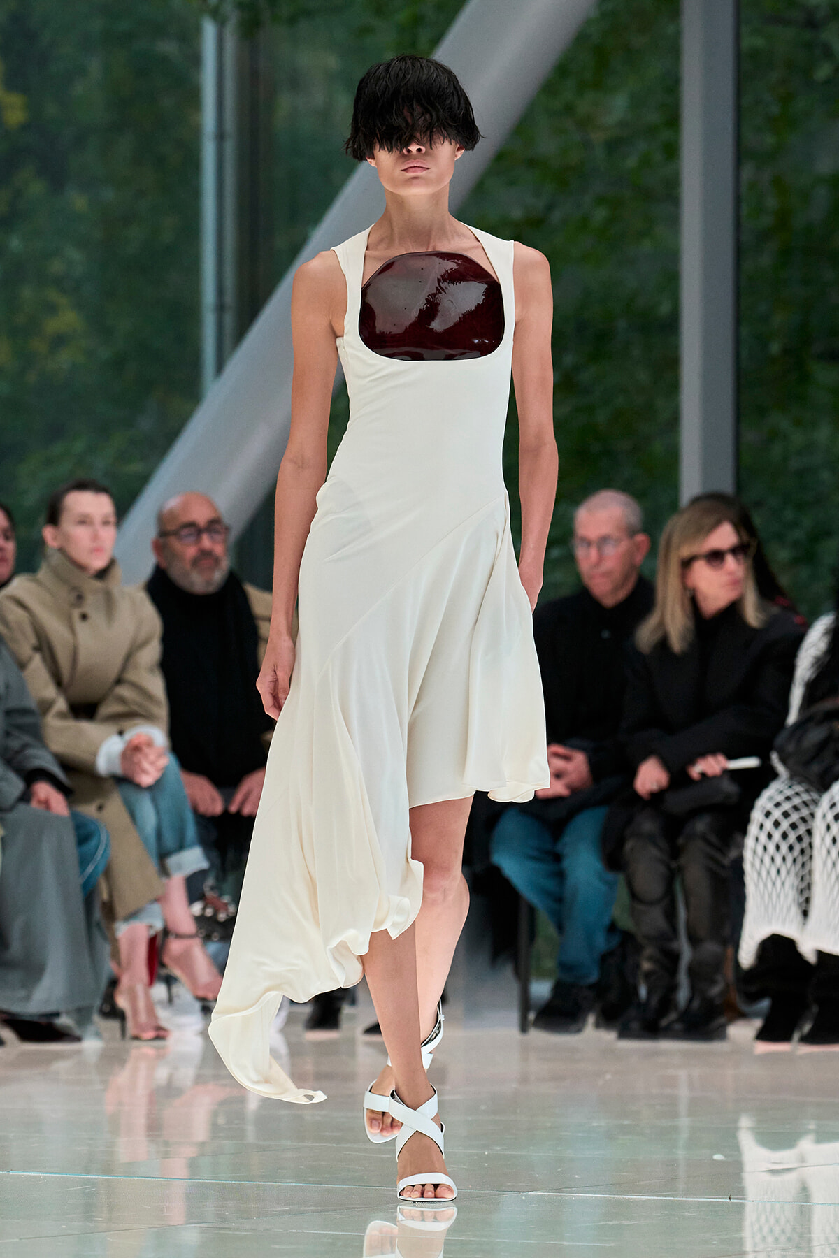 Model walking a runway in a white sleeveless dress with a glossy dark burgundy chest panel, audience in background.
