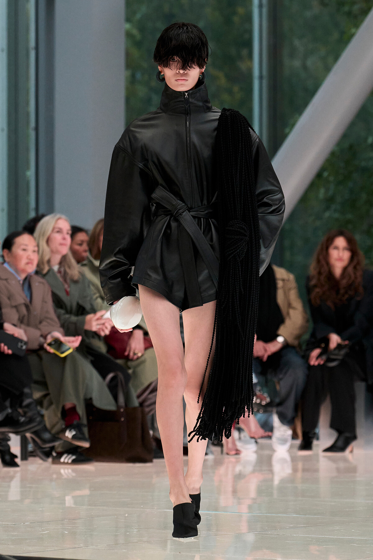 Model on a runway wearing an oversized black leather belted coat with a long black fringe scarf draped over the shoulder, carrying a white round clutch.