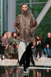 Model on a runway wearing a large snakeskin-pattern coat with a tall high collar; audience seated in the background.
