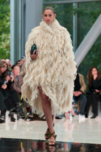 Runway model in a voluminous cream fringed coat, holding a small teal clutch, with spectators in the background.